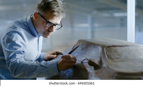 Professional Automotive Designer With Rake Sculpts Futuristic  Car Model From Plasticine Clay. He Works In A Special Studio Located In A Large Car Factory.