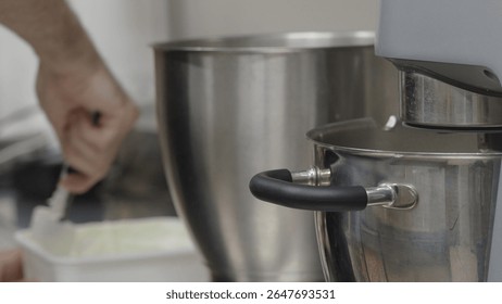 Professional baker mixing ingredients using spatula and planetary mixer, working intently in stainless steel commercial kitchen environment, revealing culinary preparation process