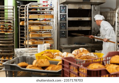 Professional baker taking out freshly baked hot bread from oven in small bakery..