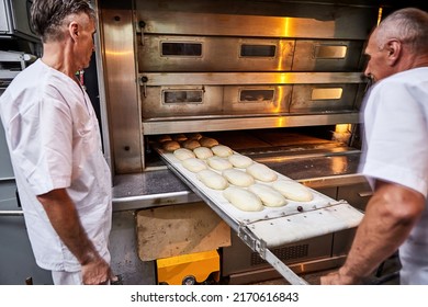 Professional Baker In Uniform Inserts Cart With Decks For Baking Raw Dough To Make Bread In An Industrial Oven In A Bakery