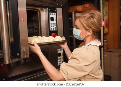 Professional Baker Wearing Medical Face Mask, Putting Tray Of Croissants Into The Oven