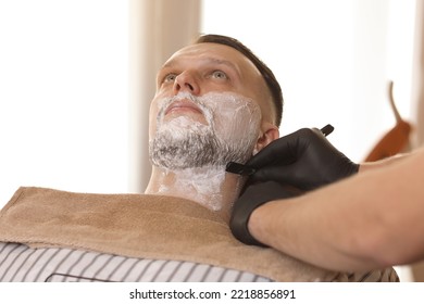 Professional Barber Shaves Customer Beard With Straight Razor. Beard Cut With Old-fashioned Blade At Barbershop. Handsome Young Man Getting His Face Shaved In Hair Studio