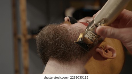 Professional barber trimming man's client beard in barbershop, close-up view. Cutting beard using hair clipper, electric trimmer machine. Hairdresser carefully cuts beard. Beauty hair salon for men. - Powered by Shutterstock