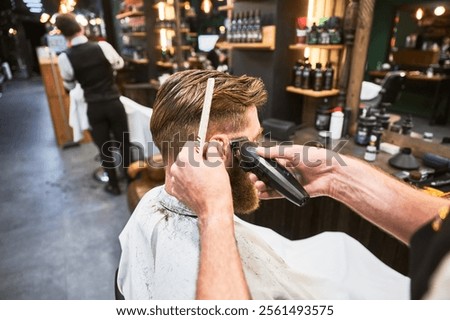 Professional barber using comb and electric shaver to cut side hair. Close-up view of male customer getting stylish haircut. Side view of man on background of other hairdresser.