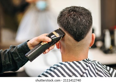 Professional barber using an electric shaver or trimmer to neaten the modern V-shaped hairstyle of a male customer viewed from the rear in close up in a barbershop - Shutterstock ID 2111412740