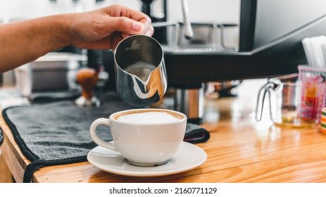 The professional barista pours the milk in the stainless steel mug into the hot coffee cup to make a cappuccino or latte in the coffee shop. how to make cappuccino coffee