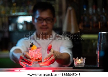 Professional barman holding decorate mixed alcoholic drink in classic glass on bar counter for serving to customer at nightclub party. Male mixologist bartender preparing liquor cocktail drink at bar