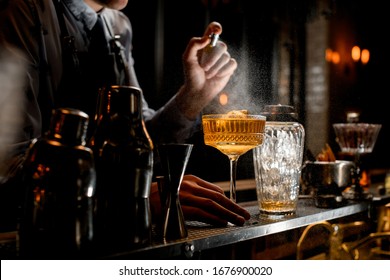 Professional Bartender Spray On Cold Brown Alcoholic Drink On Bar Counter. Nearby Stands Several Shakers.