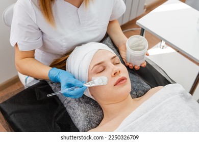 Professional beautician in salon provides comprehensive facial skin care for young woman. Hands of cosmetology specialist applying gold facial mask using brush, making skin hydrated and face glowing. - Shutterstock ID 2250326709