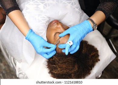 Professional Beautician woman applying Tottoo, Brow Microblading to Black african customer eyebrows to make permanent color makeup and razor blade look real in beauty salon, copy space for text logo