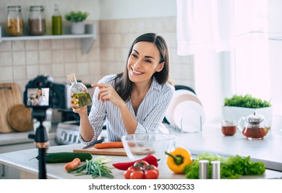 Professional beautiful happy young woman is blogging for her  kitchen channel about healthy living in the kitchen of her home and looking on camera on a tripod