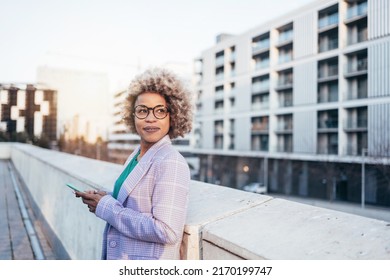 Professional Black Business Woman With Blond Hair And Trendy Eyeglasses Standing In The City Looking Away At Sunset