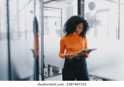 Professional Black businesswoman using tablet in modern office setting, focused and serious, wearing casual orange sweater and jeans, exemplifying mobile business management. - Powered by Shutterstock