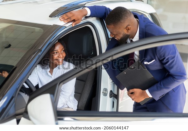 Professional Black Manager In Auto Showroom\
Consulting Female Client Sitting In Car, Young African American\
Female Choosing New Automobile In Dealership Center, Testing\
Different Vehicles
