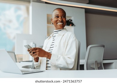 Professional black woman with a bald hairstyle sits at a desk in her office. She is smiling, holding a smartphone, and texting a message. The entrepreneur is focused on her work and uses her laptop. - Powered by Shutterstock