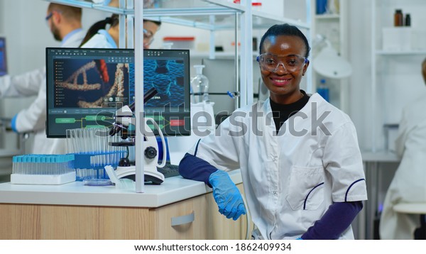 Professional black woman scientist looking\
at camera smiling in modern equipped lab. Multiethnic team\
examining virus evolution using high tech and tools for scientific\
research, vaccine\
development.