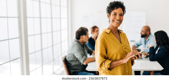 Professional black woman using a smart phone in an office. Happy business woman smiling at the camera in a boardroom. Colleagues having a meeting in the background. - Powered by Shutterstock