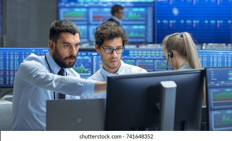 Professional Broker Consults Stock Exchange Trader At His Workstation. Multi-Ethnic Team At Stock Exchange Office Is Busy Selling And Buying Stocks On The Market. Displays Show Relevant Data Numbers.