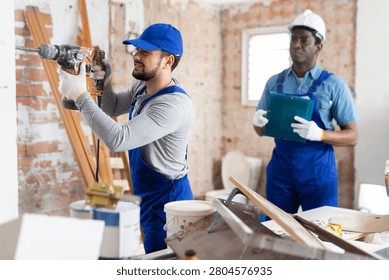 Professional builder working on a construction site indoors drills a wall with a hammer drill, and his colleague makes ..important notes in the estimate