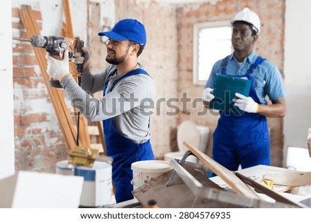 Professional builder working on a construction site indoors drills a wall with a hammer drill, and his colleague makes ..important notes in the estimate