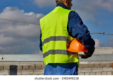 Professional Builder In Workwear Is Holding An Orange Hard Hat While Standing On The Roof Of Building Under Construction And Looking Away. Blue Sky