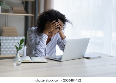 Professional burnout. Depressed upset mixed race female sit at workplace by computer hug head feel unmotivated sick lack of sleep. Desperate young black woman having problems at work missed deadline - Shutterstock ID 2036484452