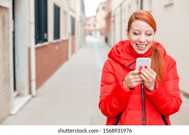 Professional Business Woman On Smartphone Smiling Happy Wearing Red Coat Outwear. Young Happy Casual Woman Texting On Smart Phone Walking Outdoor In Fall Or Winter On A Italian Street. Caucasian Model