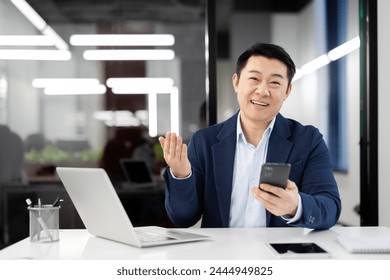 A professional businessman in a blue suit multitasking with a smartphone and laptop in a well-lit contemporary office setting. - Powered by Shutterstock