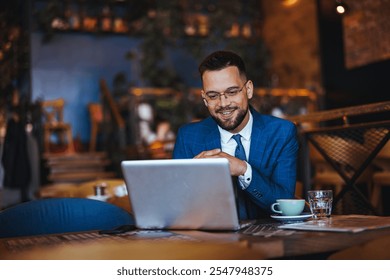 A professional businessman in a blue suit works on his laptop while smiling in a cozy cafe setting. He appears engaged and content while sipping a drink. - Powered by Shutterstock