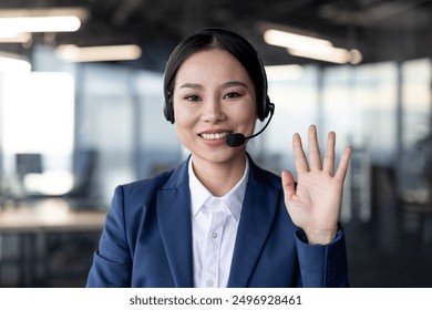 Professional businesswoman smiling at camera, wearing a headset, waving hand, and participating in video call from modern office setting. Concept customer service, remote work, virtual communication - Powered by Shutterstock