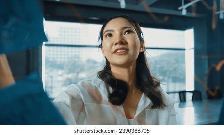 Professional businesswoman writing marketing idea on glass board. Smiling young beautiful project manager looking at colorful sticky notes with show creativity marketing strategy. Manipulator.