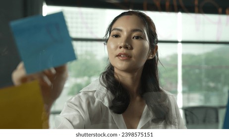 Professional businesswoman writing marketing idea on glass board. Smiling young beautiful project manager looking at colorful sticky notes with show creativity marketing strategy. Manipulator.