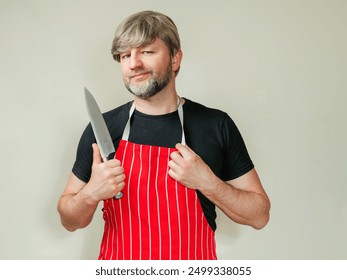 Professional butcher man in a classic white and red stripe apron holding sharp knife. Light color background. Portrait of a male in a uniform for work. He is wearing a black shirt. - Powered by Shutterstock