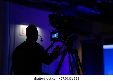 Professional camera operator filming a live event on a large broadcast television camera mounted on a tripod in a dimly lit studio with blue ambient lighting