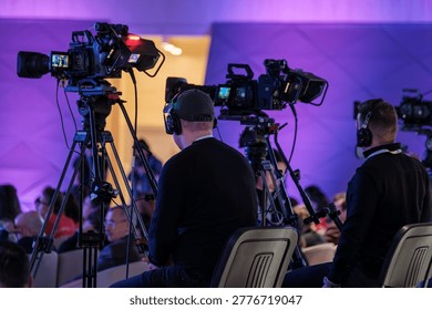 Professional camera operators capturing live event with broadcast video cameras, headsets, tripods, and purple stage lighting.