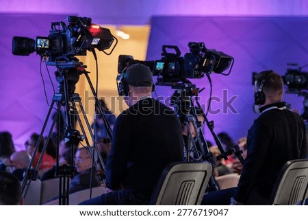 Professional camera operators capturing live event with broadcast video cameras, headsets, tripods, and purple stage lighting.