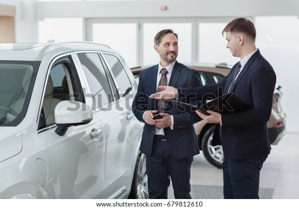 Professional car dealer showing a new car to his\
client. Mature businessman choosing a vehicle to buy communication\
occupation job service customer consumer retail retailer sales\
offering discount