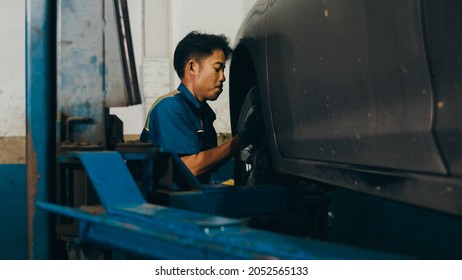 Professional Car Mechanic Changing A Car Tire On Lifted Automobile At Repair Service Station. Wheel Alignment Work At Workshop Night. Skillful Asian Guy In Uniform Fixing Car.