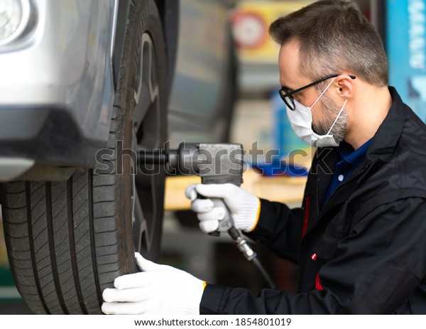 Professional car mechanic changing\
car wheel at Car maintenance and auto service garage. Caucasian man\
worker people. Surgical face mask protec coronavirus covid\
19.