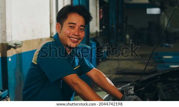 Professional\
car mechanic looking at camera and smiling at repair service\
station. Skillful Asian guy in uniform fixing car at mechanics\
garage at night. Car service\
maintenance.