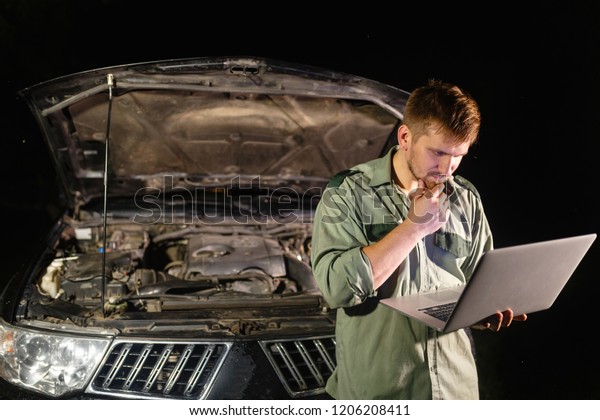 A professional car mechanic standing with\
his back to the car, working in a car service, using a laptop to\
set up and check maintenance.