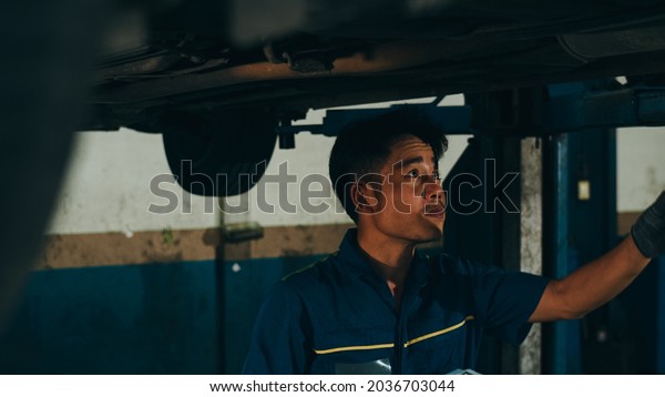 Professional car\
mechanic using paperwork makes the oil and engine check to the car\
on lifted automobile at repair service station night. Skillful\
Asian guy in uniform fixing\
car.