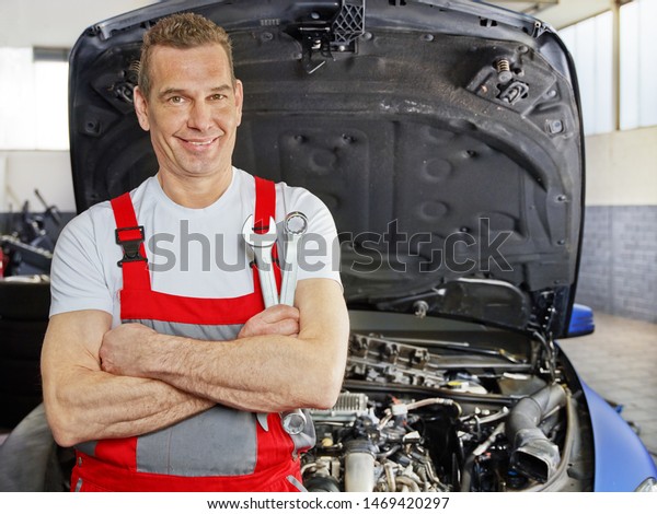 a professional car mechanical employee of a car
service workshop stand in front of a open bonnet motor engine and
hold some tools in the crossed arms and smile cheerful and friendly
in his red overall