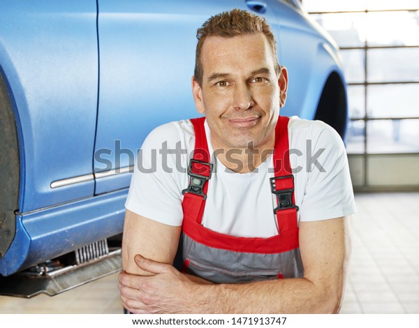 a professional car mechanical employee of a\
car and tire service workshop lean on a stack tires in front of a\
car on the hydraulic ramp and looks trusted, friendly and full of\
competition