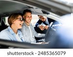 Professional Car Salesman Excitedly Showcases Sleek New Vehicle to Young Couple Inside Dealership, Hispanic Woman Holding Steering Wheel Smiling.