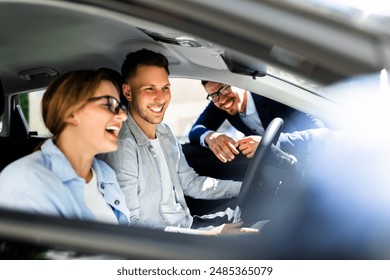 Professional Car Salesman Excitedly Showcases Sleek New Vehicle to Young Couple Inside Dealership, Hispanic Woman Holding Steering Wheel Smiling. - Powered by Shutterstock