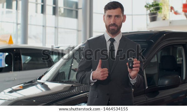 Professional car salesman\
smiling happily holding car keys standing in front of new cars at\
the dealership.