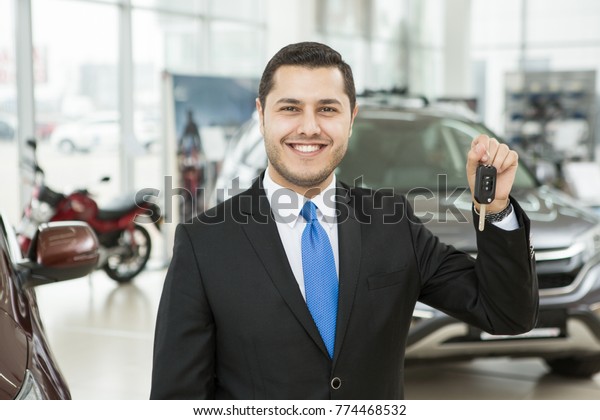 Professional car salesman smiling happily holding\
car keys standing in front of new cars at the dealership offer sale\
discount rent rental renting lease leasing selling seller retail\
retailer luxury