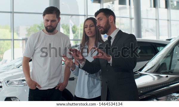 Professional car salesman is\
telling interested buyers beautiful couple about luxurious car in\
motor show while man and woman are looking at auto and listening to\
dealer.