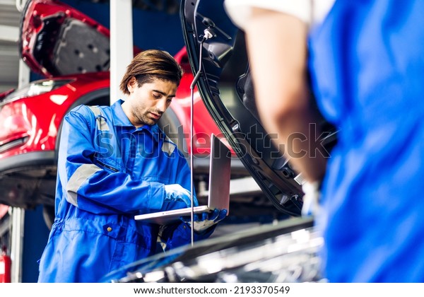 Professional car technician mechanic team in\
uniform use laptop work fixing vehicle car engine and maintenance\
repairing checking under car hood in auto service. Automobile\
service garage